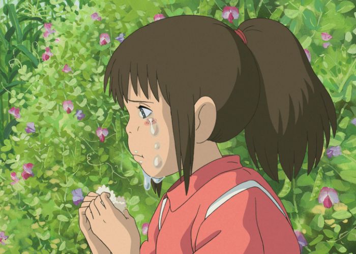 Chihiro crying in front of a green bush while eating an onigiri rice ball.